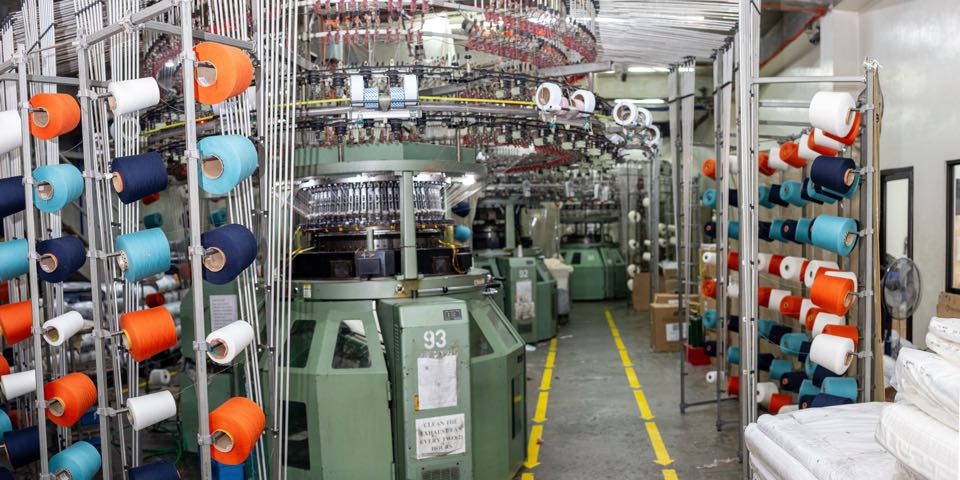 Wide view of the knitting floor: a circular knitting machine beside creels of colored yarn, with more machines along the aisle behind it.
