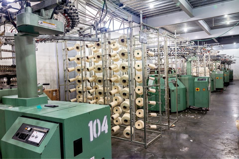 Circular knitting machine number 104 and its touchscreen control panel, with rows of yarn creels and machines behind it.
