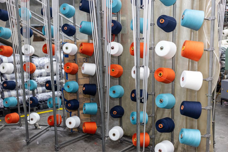 Cones of navy, orange, sky-blue and white yarn on a creel stand, threaded up to a circular knitting machine.