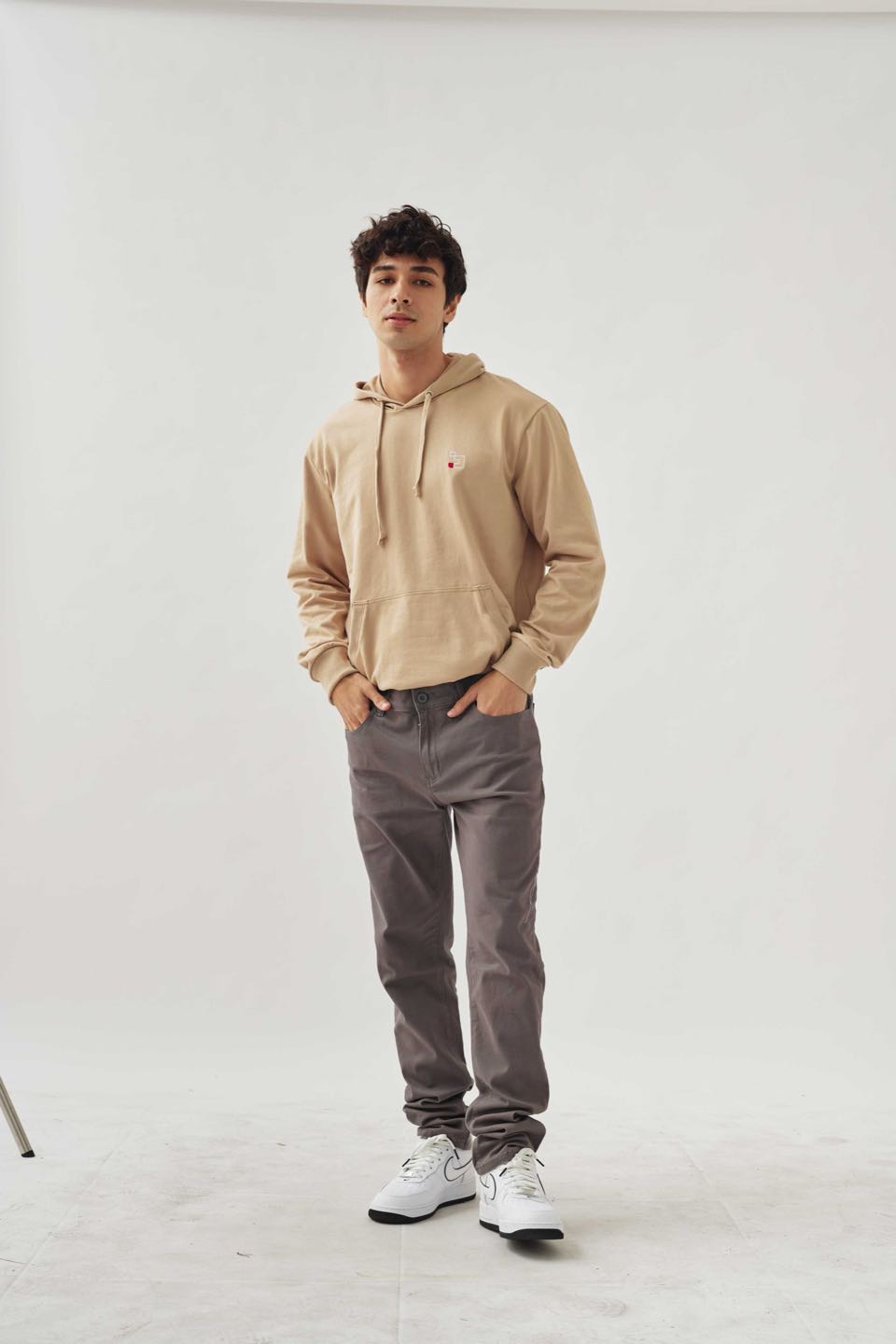 A man in a beige pullover hoodie and grey trousers standing in a white studio.