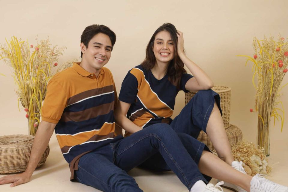 A man and a woman seated on the floor in matching mustard and navy striped knit polos.